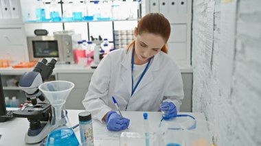 Laboratuvar önlüğü giymiş beyaz bir kadın titizlikle modern bir laboratuvar ortamında notlar alıyor..
