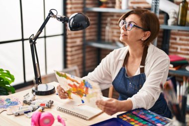 Orta yaşlı kadın gülümsüyor. Sanat stüdyosunda kendine güveni tam görünüyor.