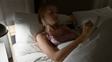 Genç sarışın kadın, yatak odasında deftere yazı yazıyor.