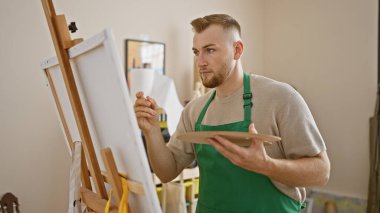 Sanat stüdyosundaki tuvale sakallı, yeşil önlük giymiş odaklanmış genç bir adam.