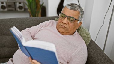 Gri saçlı ve gözlüklü son sınıf öğrencisi rahat bir oturma odasında kanepede dinlenirken kitap okuyor..