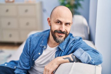 Young bald man smiling confident sitting on sofa at home
