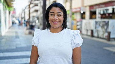 Gülümseyen orta yaşlı İspanyol kadın şehir ortamında ayakta duruyor, portre, gündüz vakti, sıradan, neşeli.