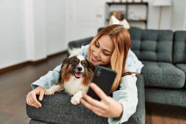Beyaz bir kadın köpeğe sarılıyor ve evde akıllı telefondan selfie çekiyor.