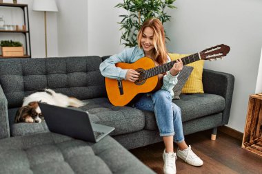 Beyaz bir kadın online klasik gitar dersi alıyor. Evde köpekle birlikte kanepede oturuyor.