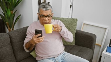 Orta yaşlı, gözlüklü, akıllı telefon kullanan ve elinde kupa tutan bir adam.