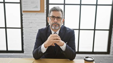 Genç Latin asıllı, gri saçlı, iş adamı ofiste ciddi bir yüzle masada oturuyor.