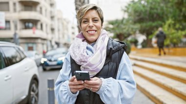 Kısa sarı saçlı, gülümseyen İspanyol kadın kalabalık bir caddede akıllı telefon kullanıyor..