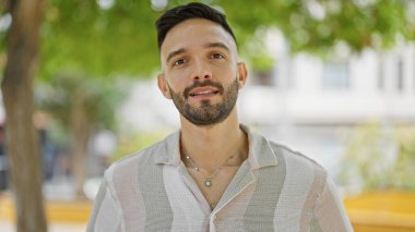 Young hispanic man standing with serious expression at park