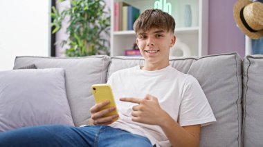 Gülümseyen genç, beyaz, sarışın bir genç, rahat bir oturma odasında akıllı telefonunu işaret ederek, modern iç mekan rahatlığını somutlaştırıyor..