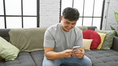 Neşeli Asyalı bir adam, modern oturma odasındaki gri bir koltukta otururken akıllı telefonuyla gelişigüzel kayıyor..