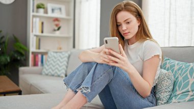 Evdeki kanepede oturan akıllı telefon kullanan genç sarışın kadın.