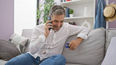 Mutlu, genç İspanyol bir adam, gri saçlı, akıllı telefondan iş konuşuyor, elinde kart, ev koltuğunda dinleniyor..