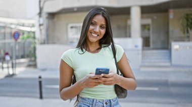 Sokakta gülümseyen akıllı telefon kullanan genç Latin kadın.