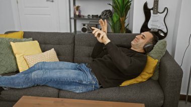 İspanyol adam gri koltukta dinleniyor, kulaklıklar takılı, akıllı telefon elinde, gitar arka planı, günlük, boş zaman, kapalı.