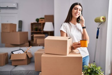 Young beautiful hispanic woman talking on smartphone drinking coffee at new home