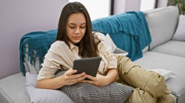 Evde teknoloji zamanı, güzel İspanyol bir kadın, rahatça dokunmatik pedini kullanıyor, rahatça oturuyor, ciddi ama rahat bir ifadeyle, rahat bir kanepede, kapalı bir oturma odasında.