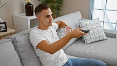 Yakışıklı adam, elinde pembe kupa ve uzaktan kumandayla gri bir koltukta rahatça oturuyor.