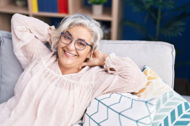 Orta yaşlı, kır saçlı, elleri başının üstünde, evinde kanepede oturan bir kadın.