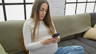 Evdeki koltukta oturan hamile bir İspanyol kadın, sakin bir oturma odasında akıllı telefonunu kullanıyor..