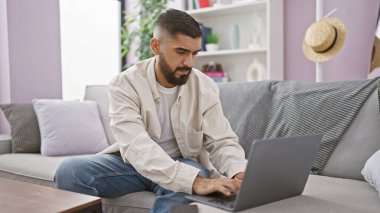 Modern bir oturma odasındaki koltukta dizüstü bilgisayar kullanan sakallı yakışıklı bir genç..