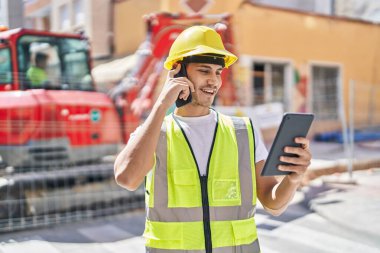 Young hispanic man architect talking on smartphone using touchpad at park