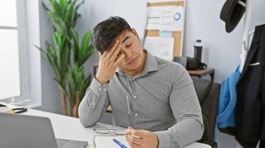 Stresli Asyalı adam, elleri alnında modern ofiste masa başında oturuyor, iş yorgunluğunu tasvir ediyor..