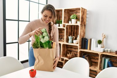 Genç, güzel, İspanyol bir kadın gülümsüyor. Elinde alışveriş torbası var.