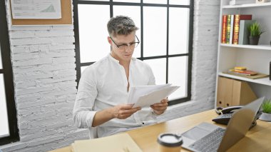 Ciddi yüzlü beyaz erkek yönetici, bir iş belgesini okumaya dalmış, çalışkan işçi kapalı ofis ortamında masa başı işinde gelişiyor..