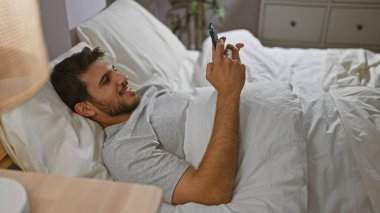 Sakallı, gülümseyen, genç bir İspanyol adam yatağında uzanıyor, akıllı telefonunu sıcacık, aydınlık bir yatak odasında tutuyor..