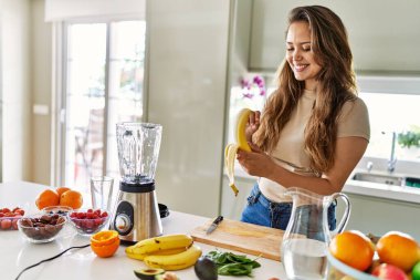 Genç, güzel İspanyol kadın mutfakta muz soyan blender ile sebze suyu hazırlıyor.