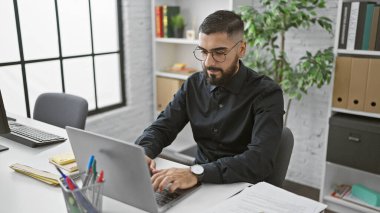 Modern ofiste dizüstü bilgisayarda çalışan sakallı bir adam şirket ortamında üretkenlik ve profesyonellik sergiliyor..