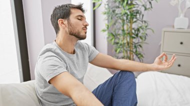 Huzurlu bir yatak odasında, arka planda bitki olan bir yatakta meditasyon yapan gevşemiş bir İspanyol..