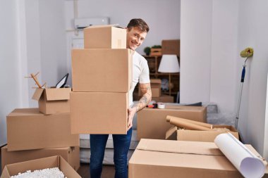 Genç adam yeni evinde paketleri tutarken kendinden emin gülümsüyor.