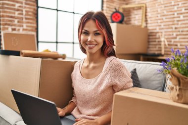 Yeni evindeki kanepede oturan beyaz bir kadın dizüstü bilgisayar kullanıyor.