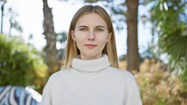 Beyaz kazak giyen, mavi gözlü çekici sarışın kadın güneşli bir parkta poz veriyor..