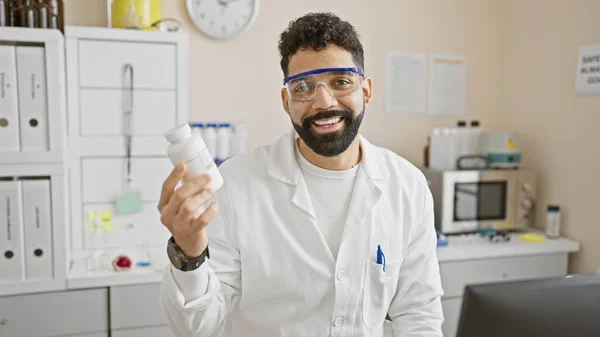 医療ボトルを持っている実験室のコートの笑顔の若いヒスパニック男性は バックグラウンドの機器を持つクリニックに立っています — ストック写真