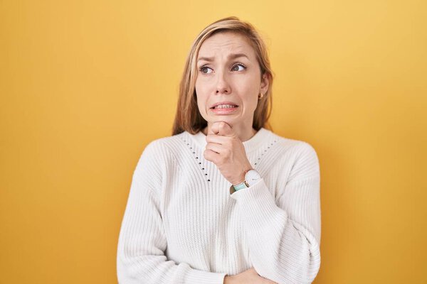 Young caucasian woman wearing white sweater over yellow background thinking worried about a question, concerned and nervous with hand on chin 