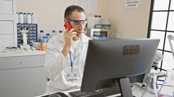 Hispanic scientist in lab coat analyzing data on computer while talking on phone in laboratory.