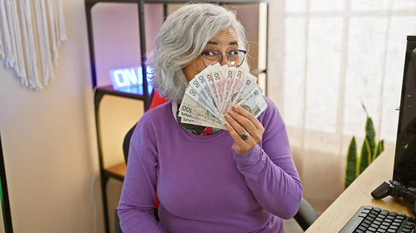 Mature woman covers mouth with polish zloty banknotes, indoor home office with gaming gear