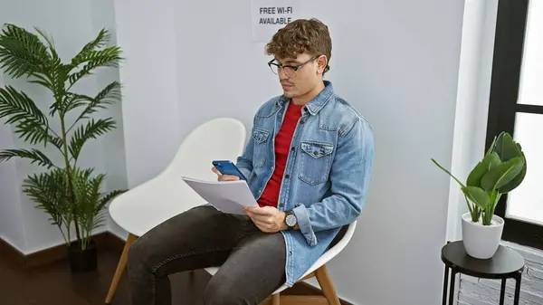Handsome, young hispanic man engrossed in a serious text conversation on his smartphone, while anxiously clutching documents and sitting in a waiting room chair, indoor portrait.