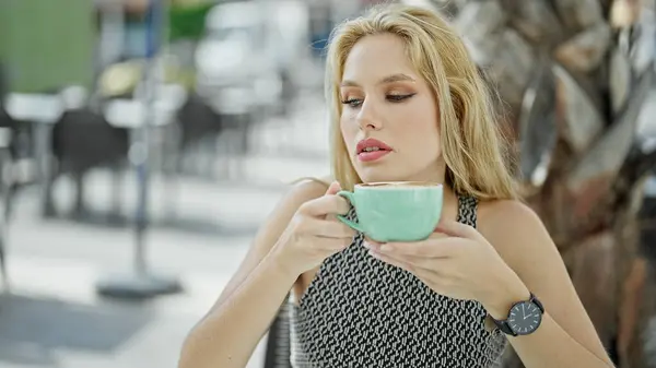 Young blonde woman holding cup of coffee sitting on table at coffee shop terrace
