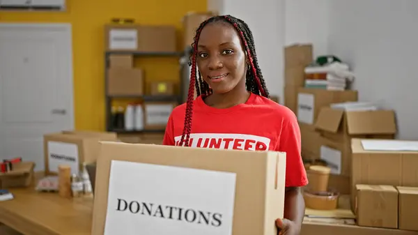 Beautiful african american woman with braids, confidently smiling while volunteering at charity center, holding donations package in the warehouse