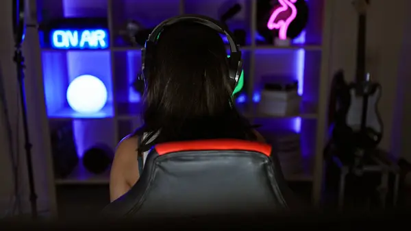 Backwards-sitting young, beautiful hispanic woman streamer brings the heat in nighttime gaming room stream, surrounded by the glow of gaming tech and the rhythm of the game\'s beat.