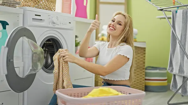 若い金髪の女性は自信を持って洗濯室で親指でジェスチャーをしている服を洗う — ストック写真