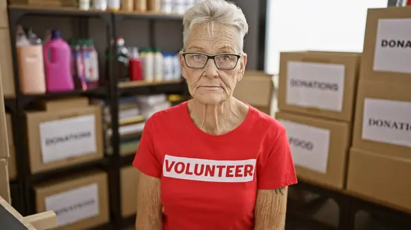 Senior grey-haired lady sitting at the charity center table, offering volunteer services with a serious face.