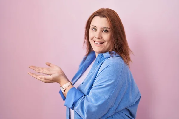 Young hispanic woman with red hair standing over pink background pointing aside with hands open palms showing copy space, presenting advertisement smiling excited happy