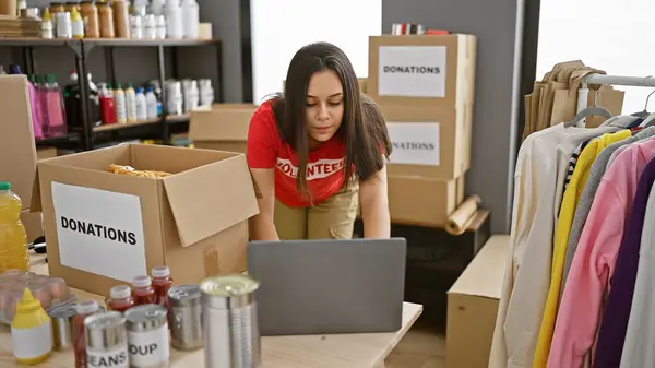 Portrait of a dedicated, young, beautiful hispanic woman volunteer working tirelessly over laptop in a charity center, providing online support and donations amidst boxes in warehouse