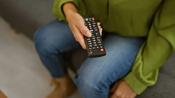A latina woman casually dressed in jeans and a sweater holding a remote control in a cozy living room setting.