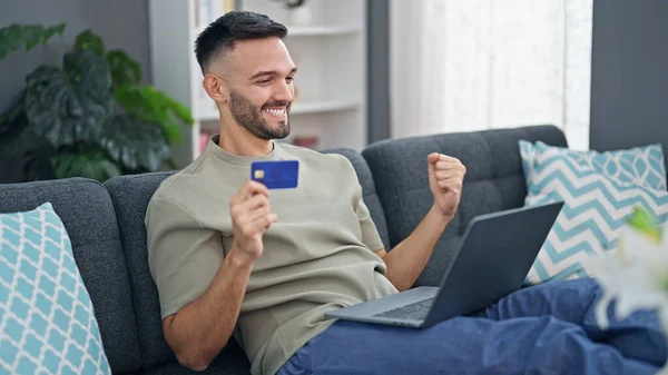 Young hispanic man shopping with laptop and credit card with winner gesture at home